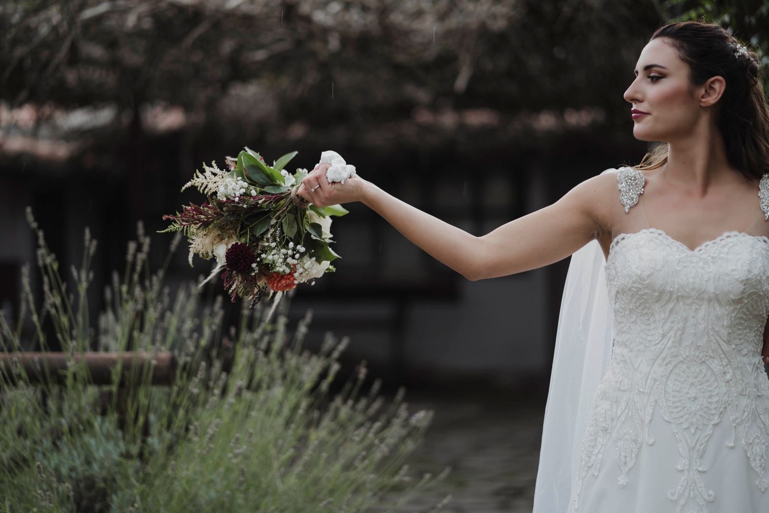 Bride in white lace dress holds out bouquet with greenery and white flowers against garden backdrop.