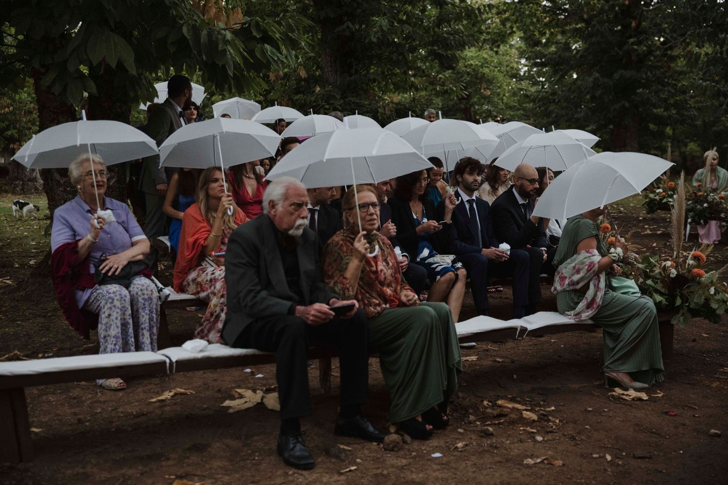 Wedding guests seated on benches holding white umbrellas during outdoor ceremony.