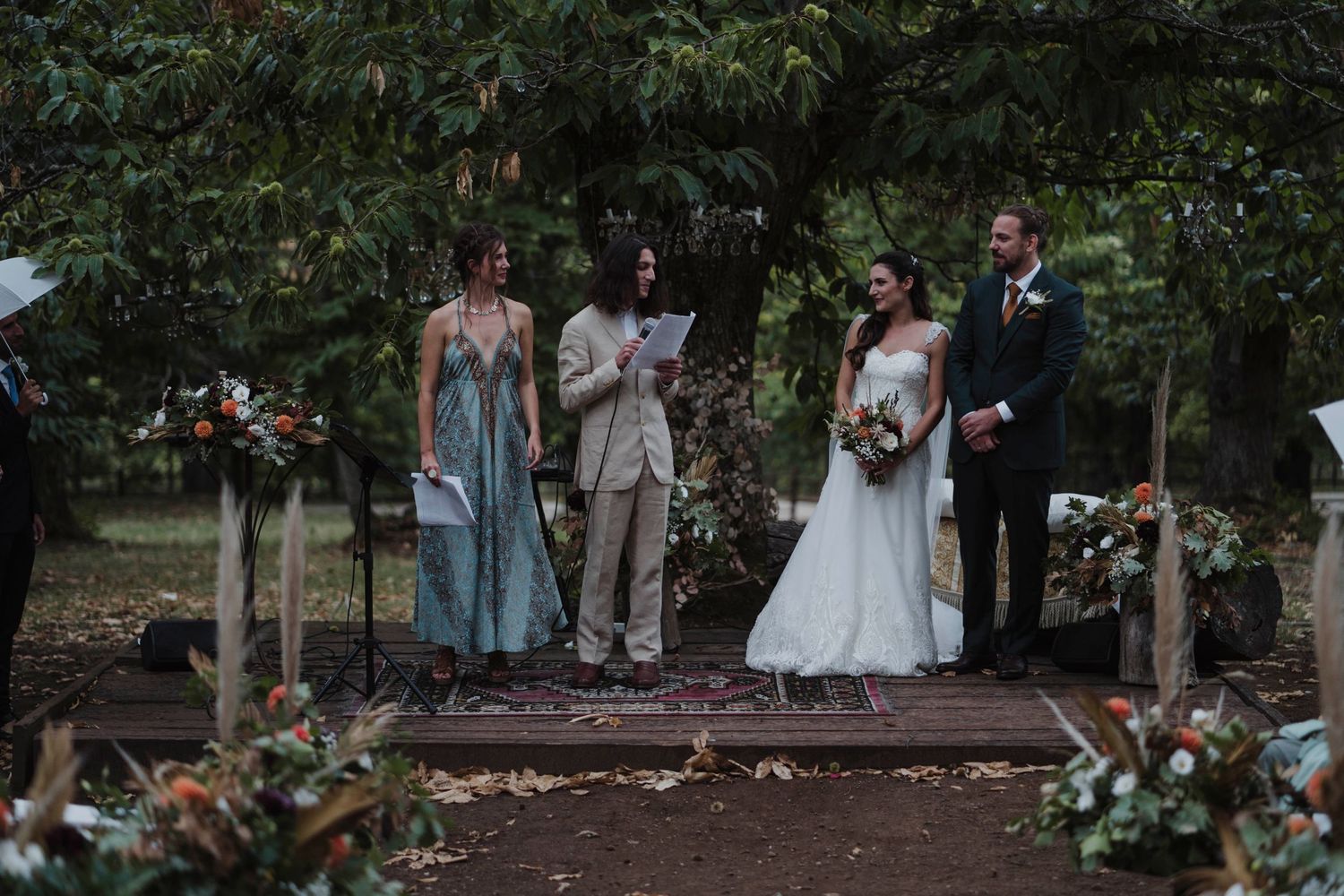 Wedding party stands on elevated wooden platform during outdoor forest ceremony.