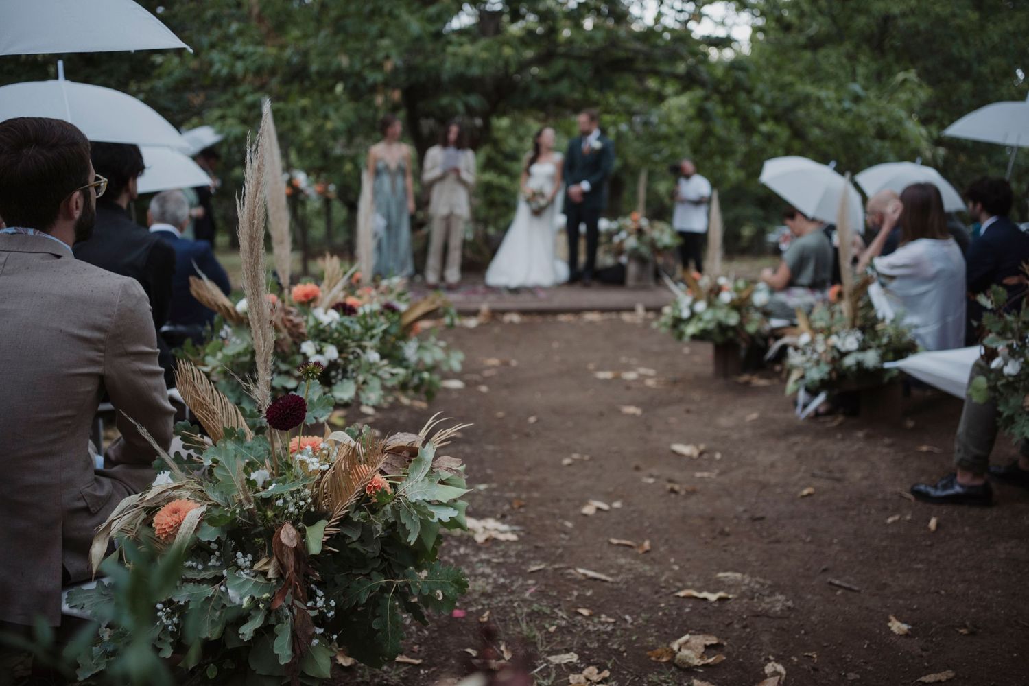 Outdoor wedding ceremony on a dirt path with white umbrellas and floral decorations along the aisle.