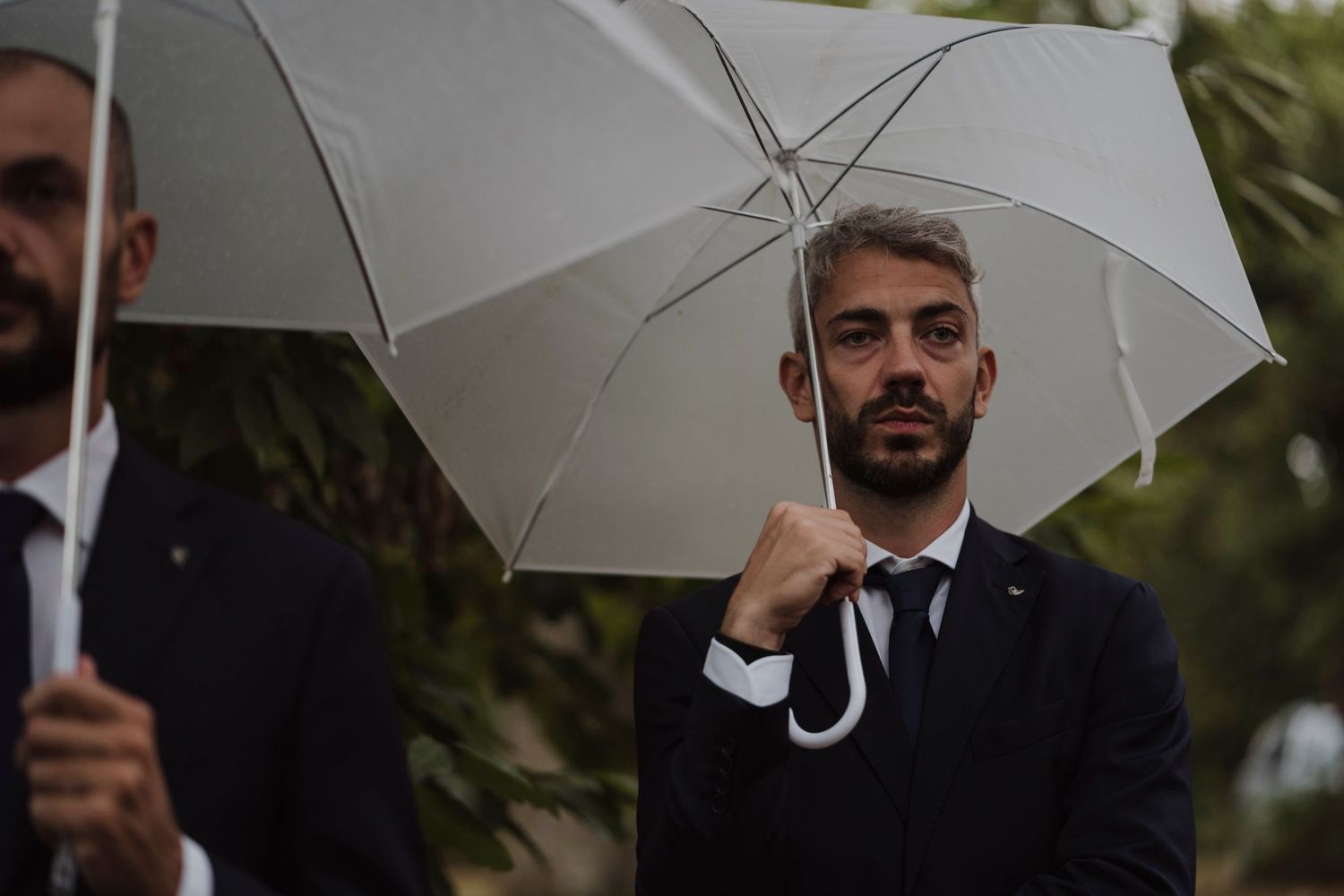 Wedding guest in formal attire holds white umbrella during outdoor ceremony.