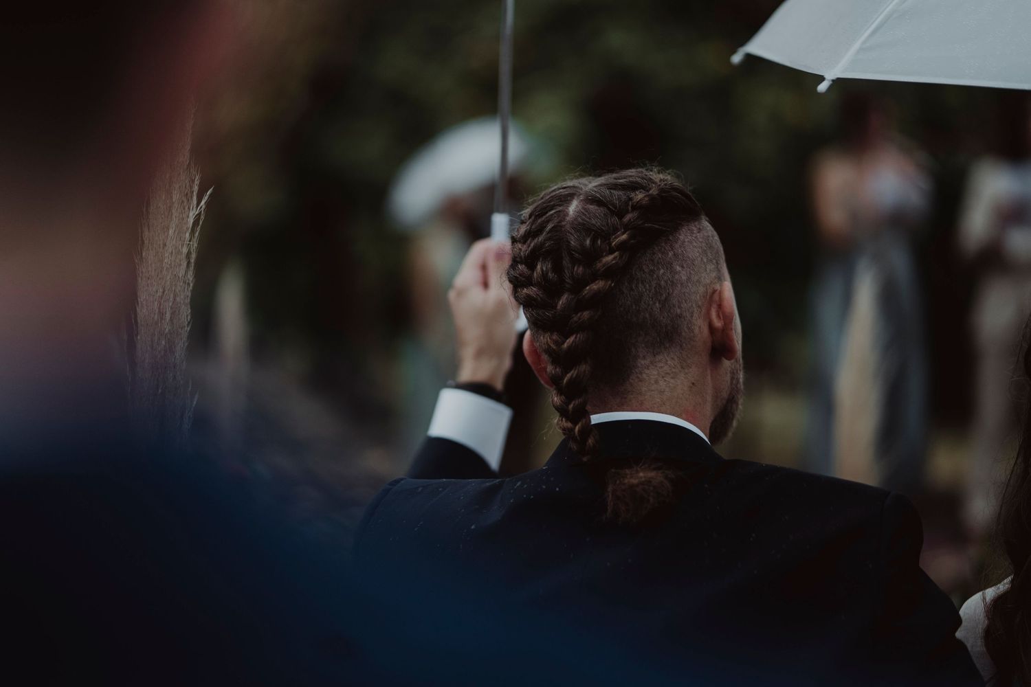 A formal wedding guest in black attire holds a white umbrella during an outdoor ceremony.