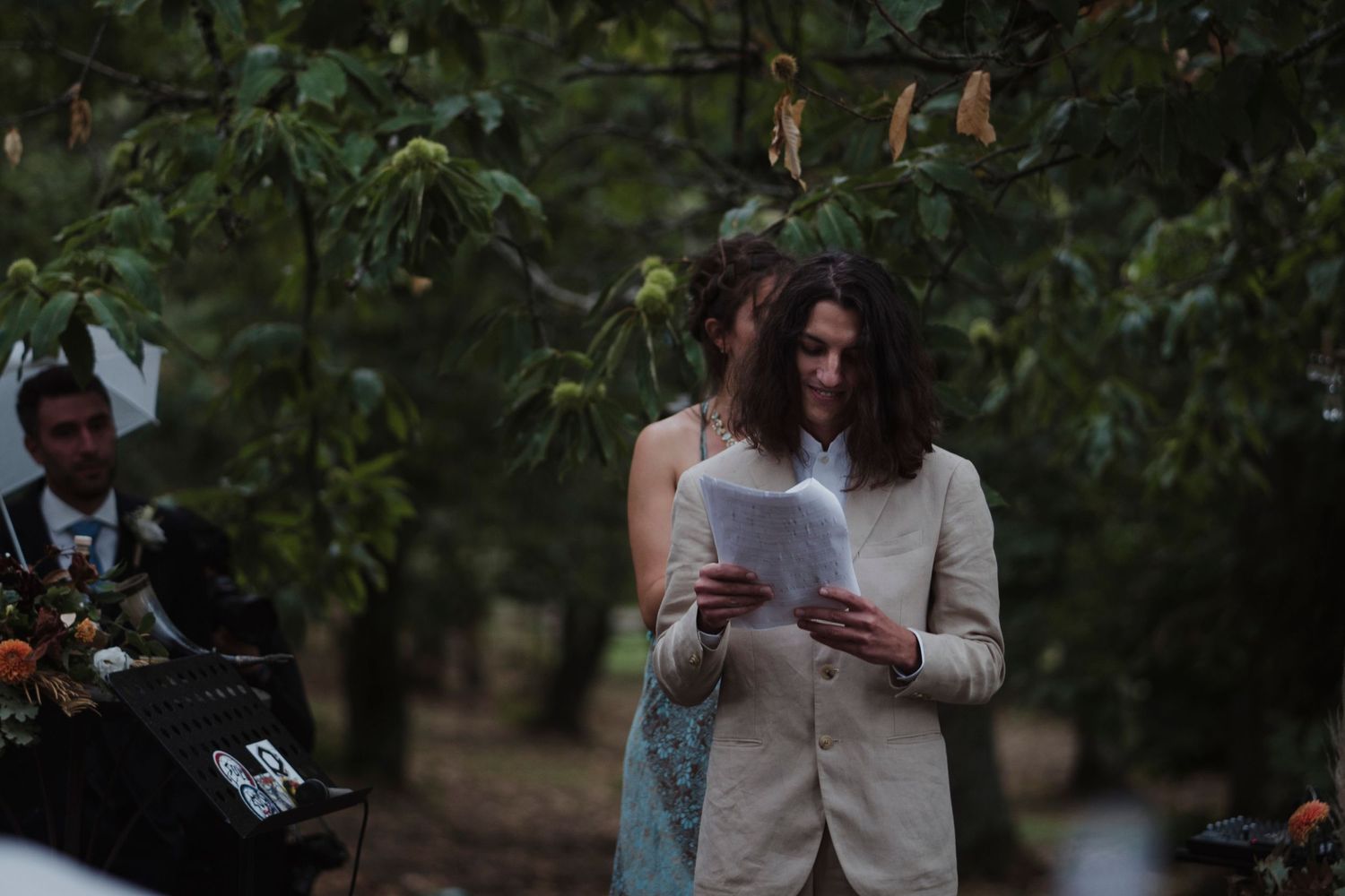 Guest reads from paper during outdoor wedding ceremony surrounded by trees and natural scenery.