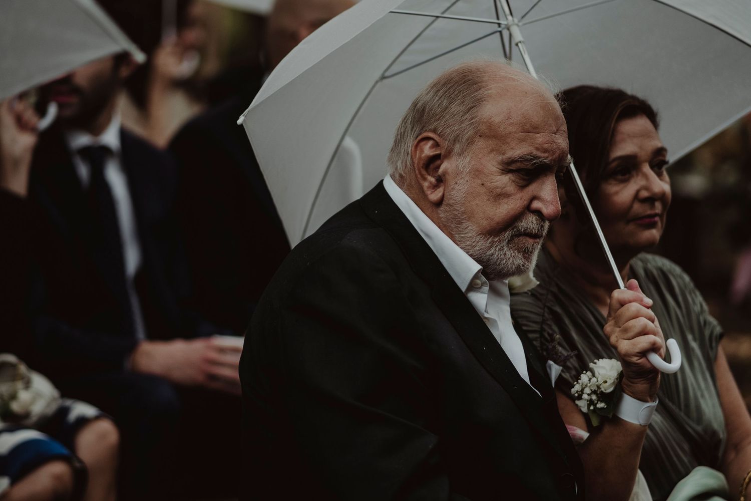 Wedding guests hold white umbrellas during an outdoor ceremony in a garden setting.