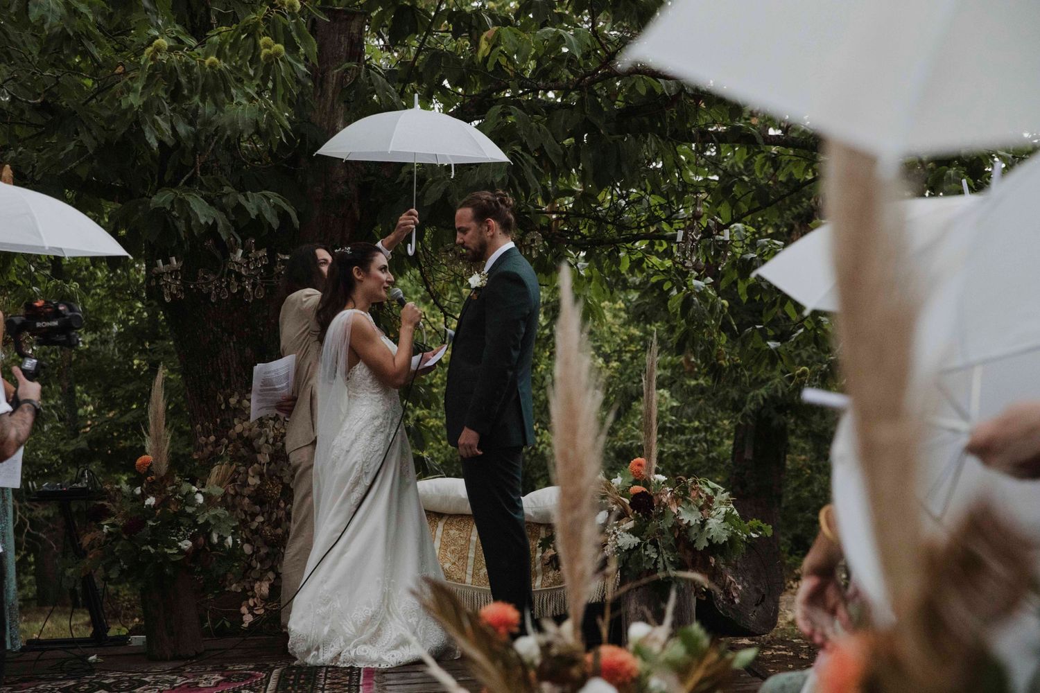 A couple exchanges vows during an outdoor wedding ceremony under white umbrellas surrounded by greenery.