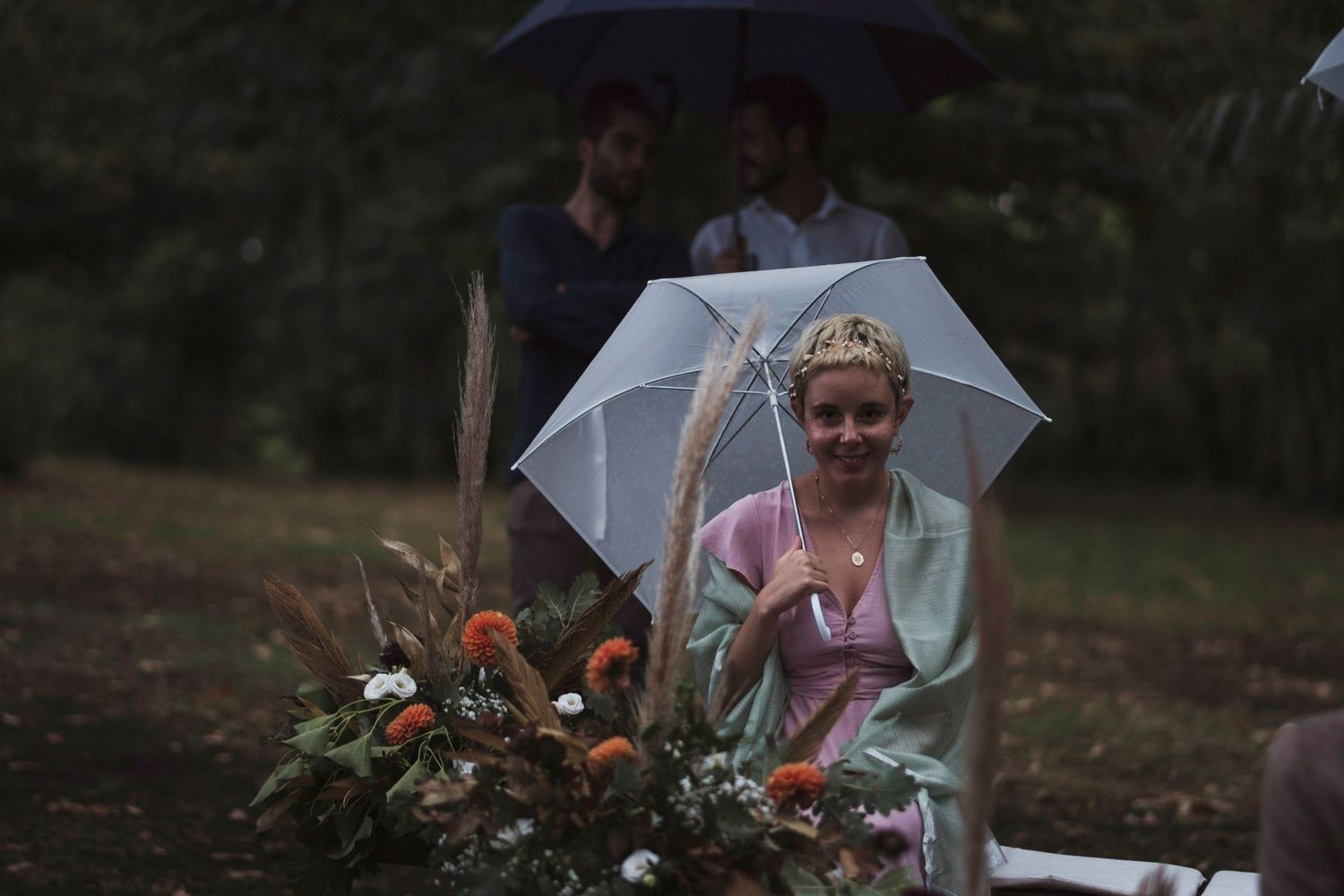 Wedding guests holding white umbrellas near floral arrangements during an outdoor ceremony.