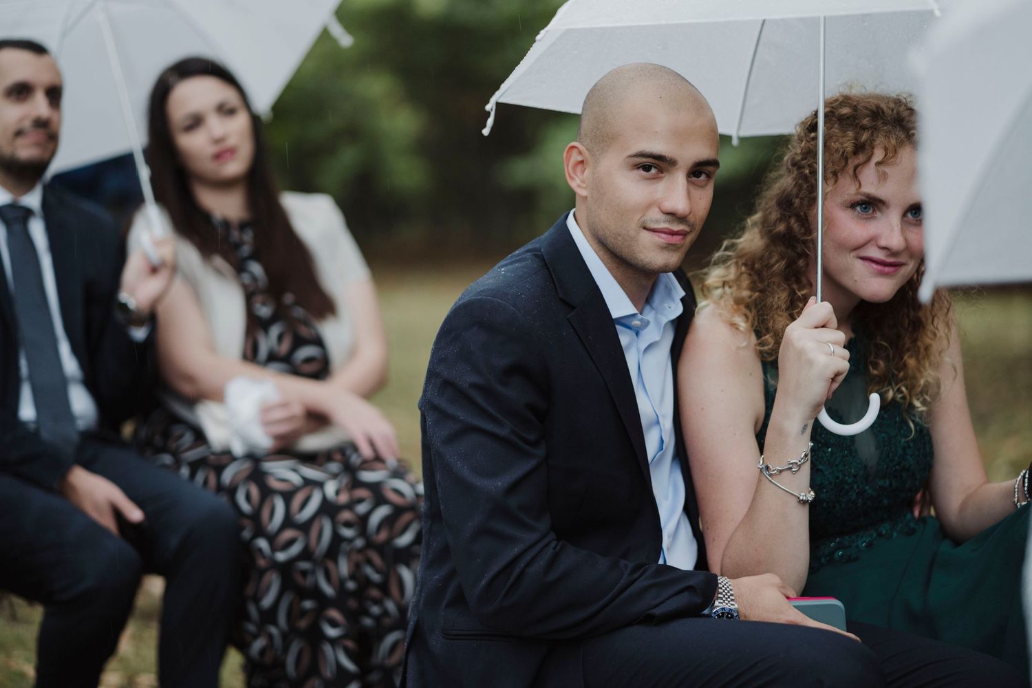 Wedding guests seated with white umbrellas at an outdoor ceremony.