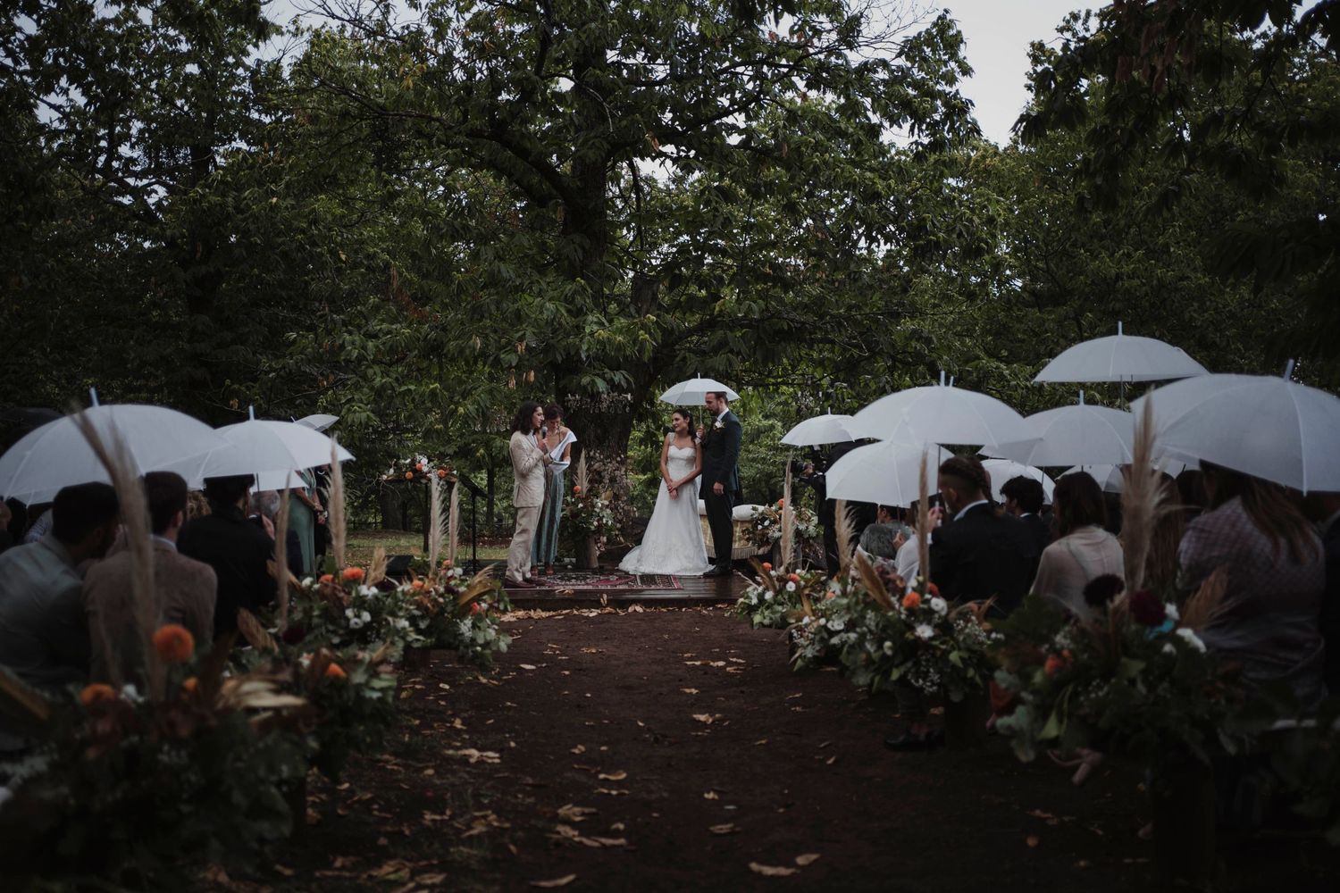 Guests with white umbrellas attend an outdoor woodland wedding ceremony.