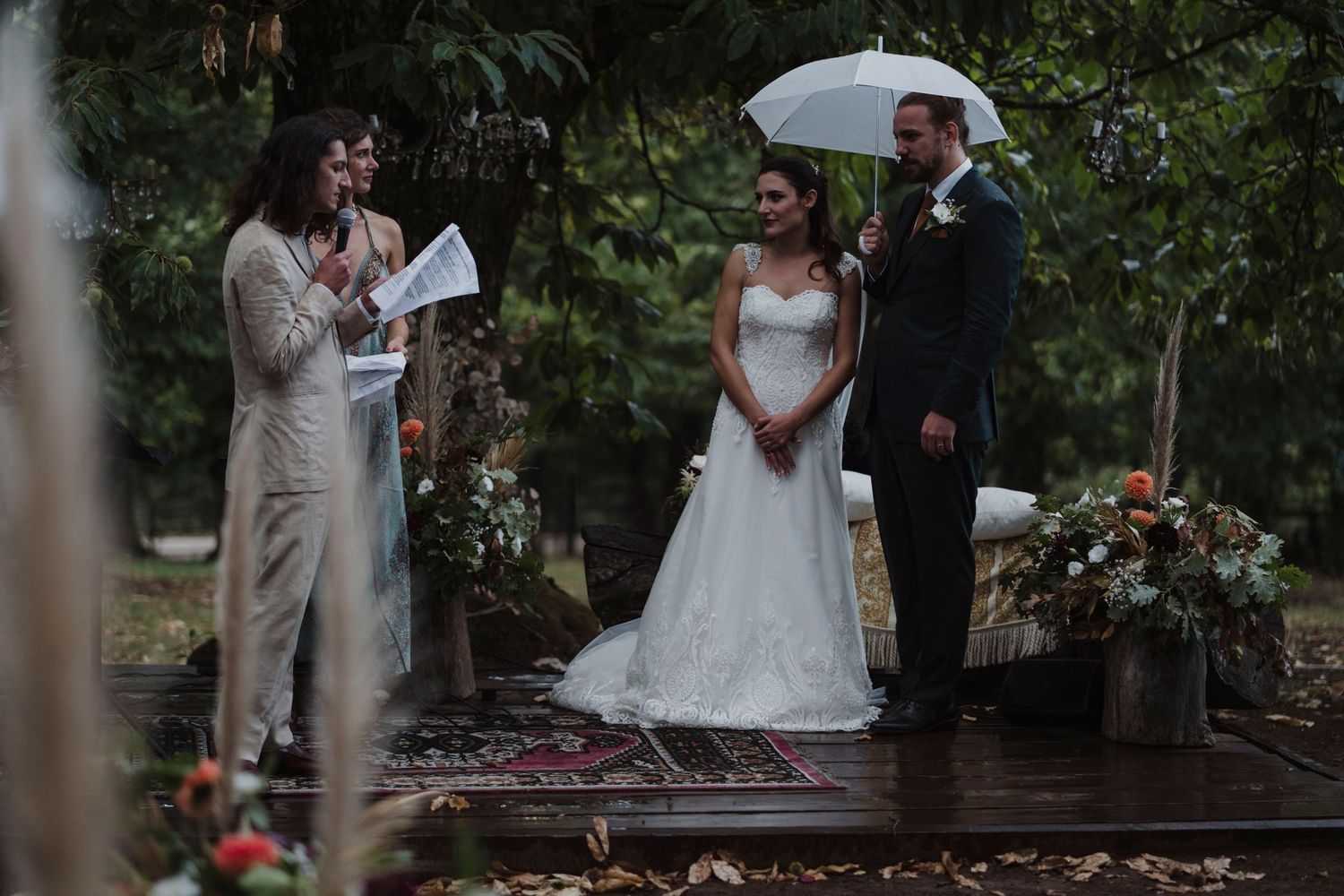 A rainy outdoor wedding ceremony with a couple and officiant under white umbrellas.
