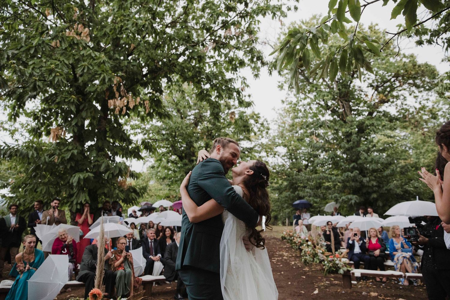 An outdoor wedding ceremony under trees with guests holding white umbrellas.