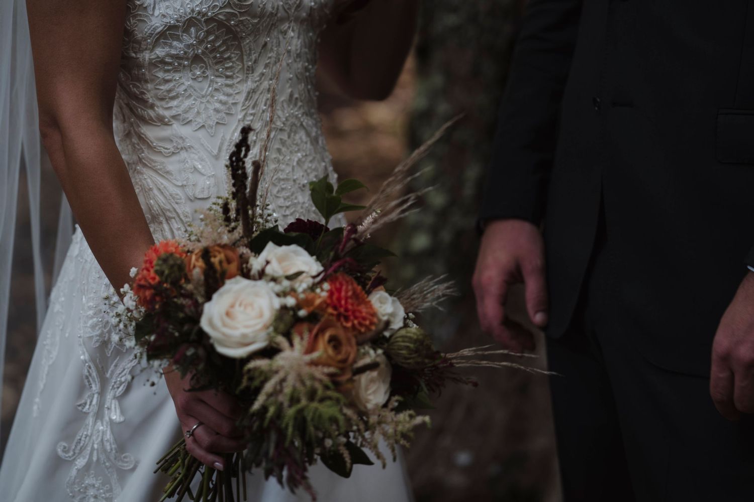 A wedding bouquet with white roses and orange accents held against a lace dress and dark suit.