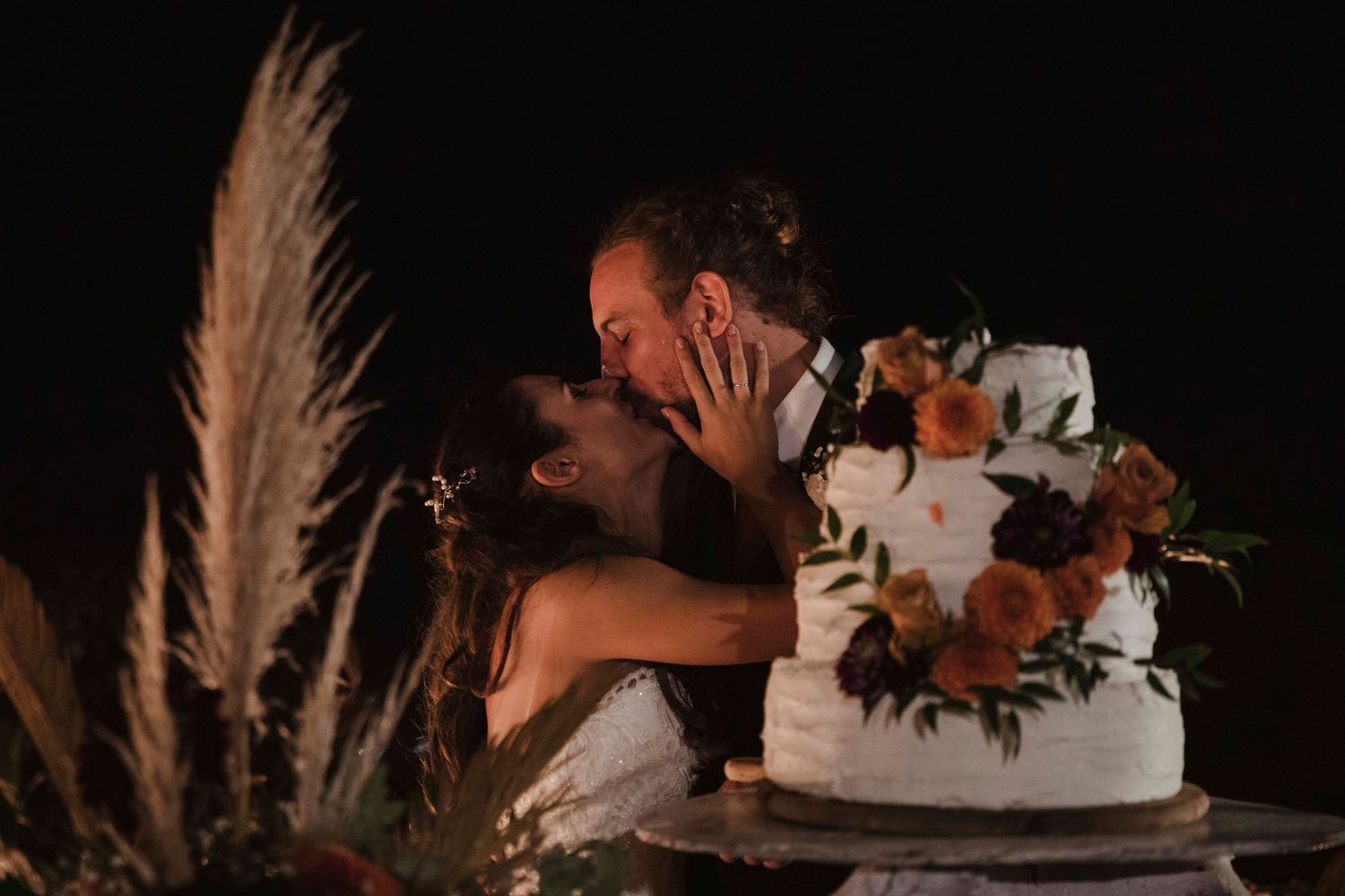Newlyweds share a passionate kiss behind their wedding cake decorated with autumn flowers.