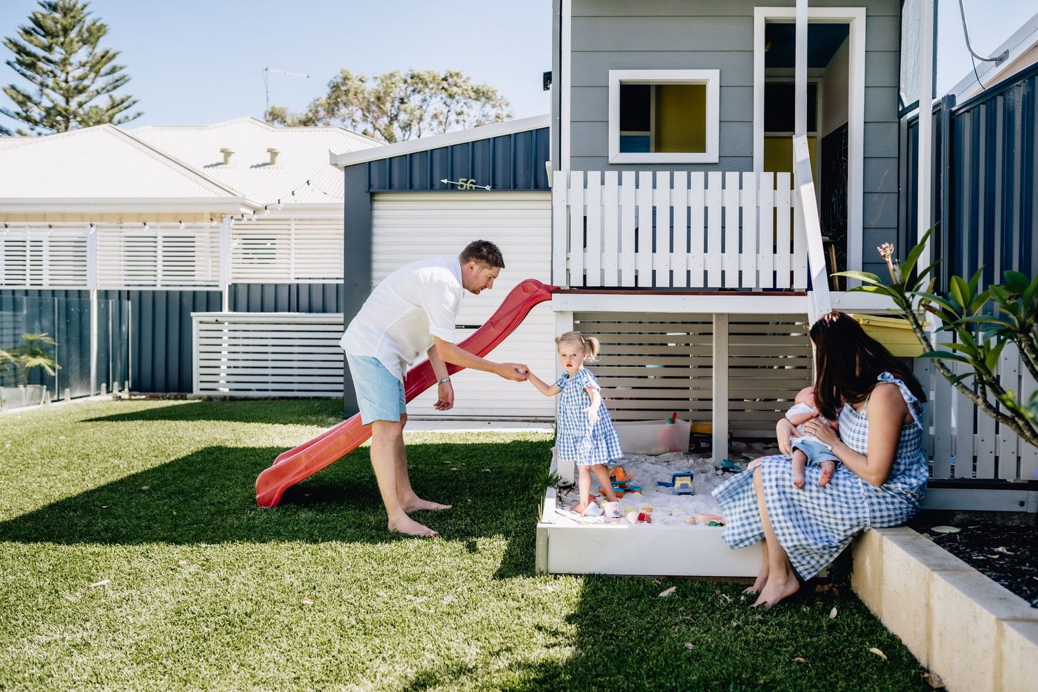 Mum, Dad, toddler and small baby playing around a cubby house in their backyard in Perth