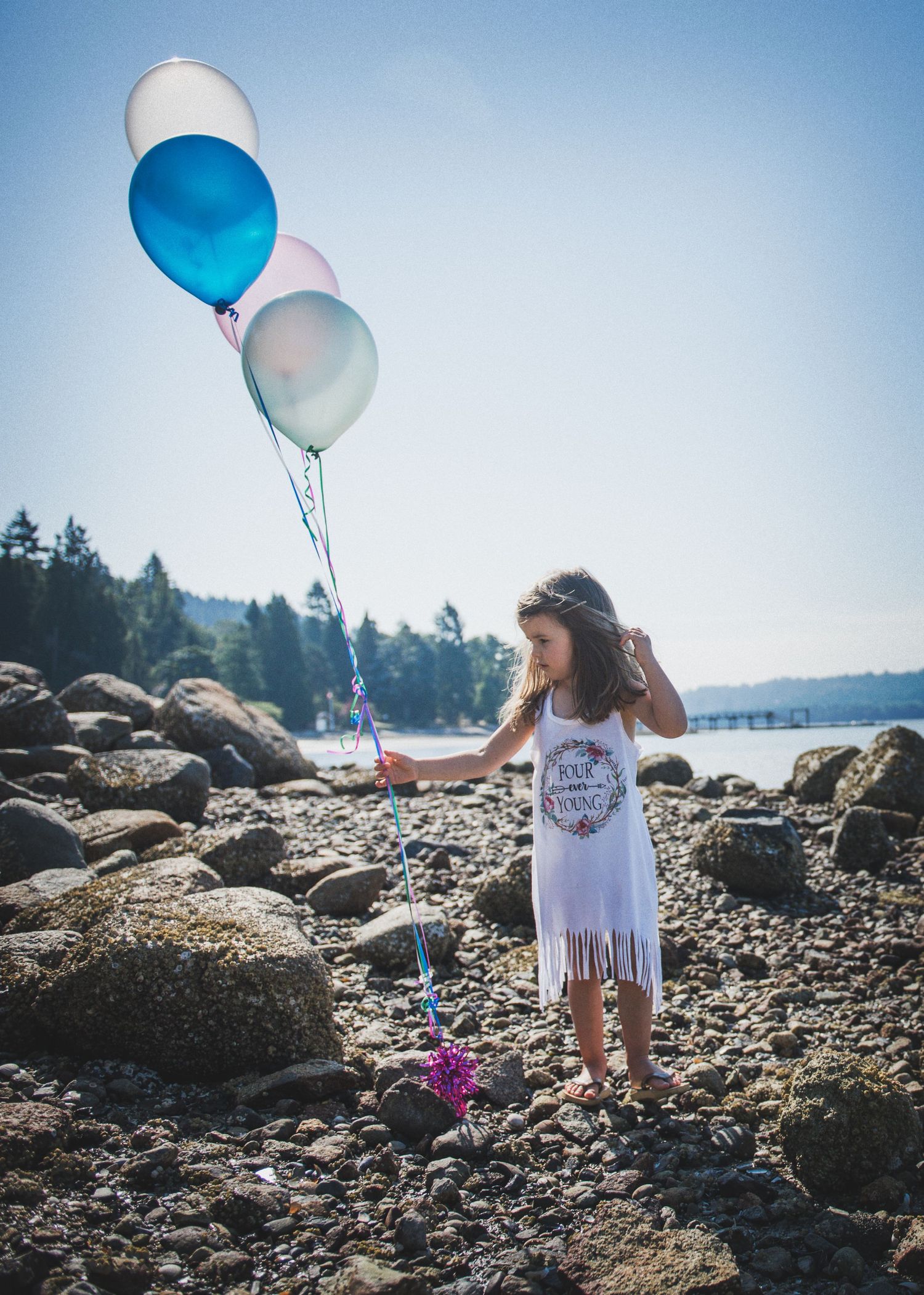 A dreamy summer beach scene with blue and white balloons against a rocky shoreline and evergreen trees.