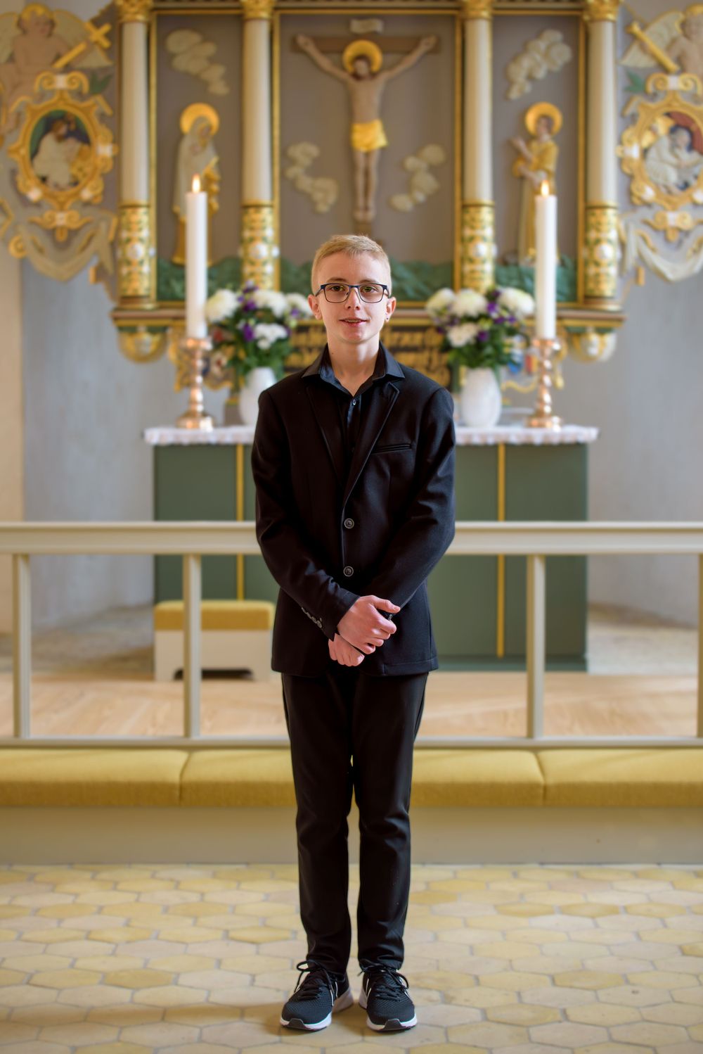 Young boy in black suit standing before ornate church altar with crucifix and floral decorations during confirmation.