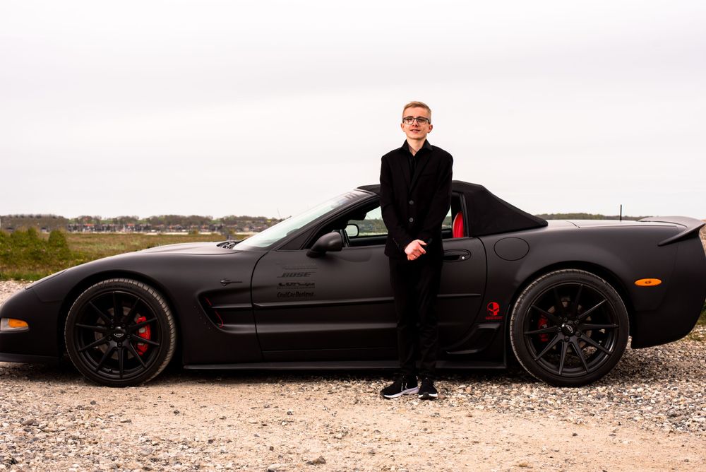 Young boy dressed in black suit standing proudly beside a matte black Corvette on a gravel surface.