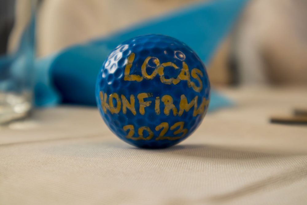Blue golf ball painted with gold text reading Lucas Konfirmation 2023 on a white tablecloth.