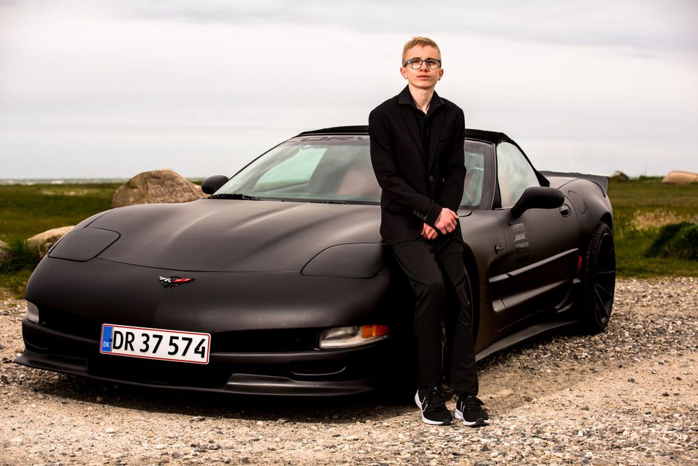 Young boy in black suit leaning against a matte black Corvette sports car outdoors in Denmark.
