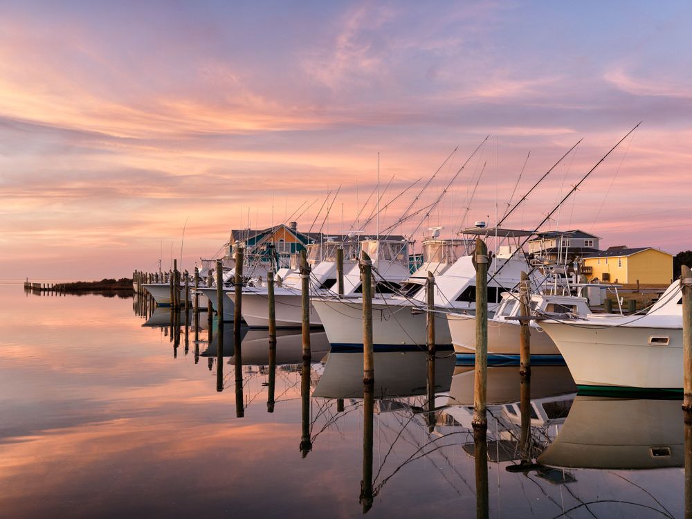 Row of fishing boats docked at marina during colorful sunset with pink sky reflections.