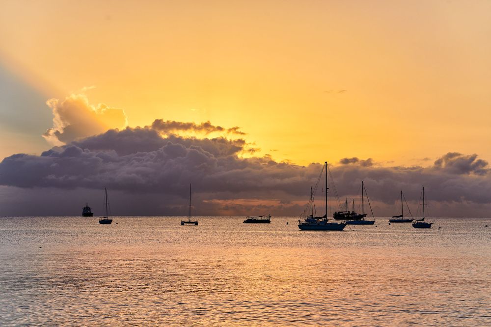 Sailboats silhouetted against a dramatic orange and purple sunset over calm ocean waters.