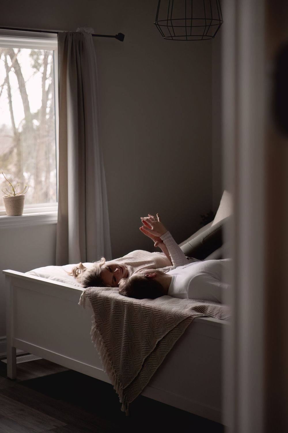 A person relaxes in bed beside a window, holding a phone in the soft morning light.