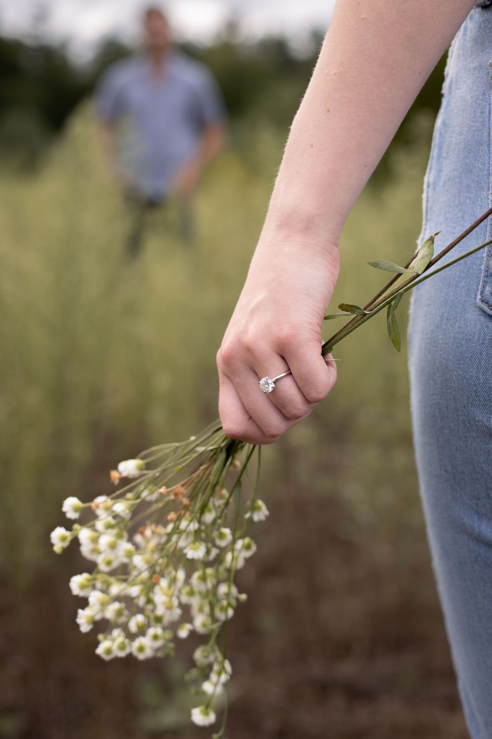Hand holding delicate white wildflowers while wearing jeans, with engagement ring visible.