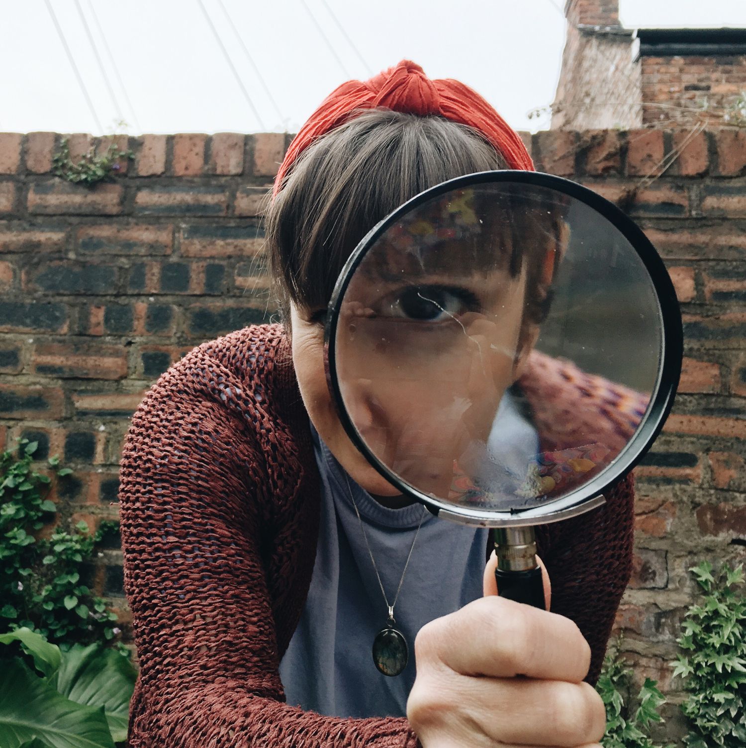 Someone peers through a large magnifying glass against a brick wall background while wearing a red headband.