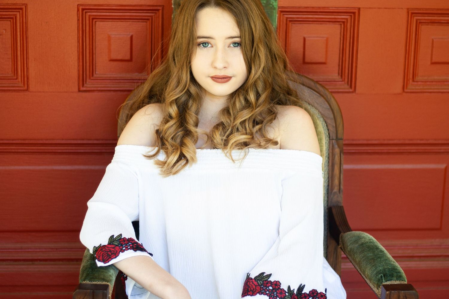 A person in a white off-shoulder top poses against a red wall background.