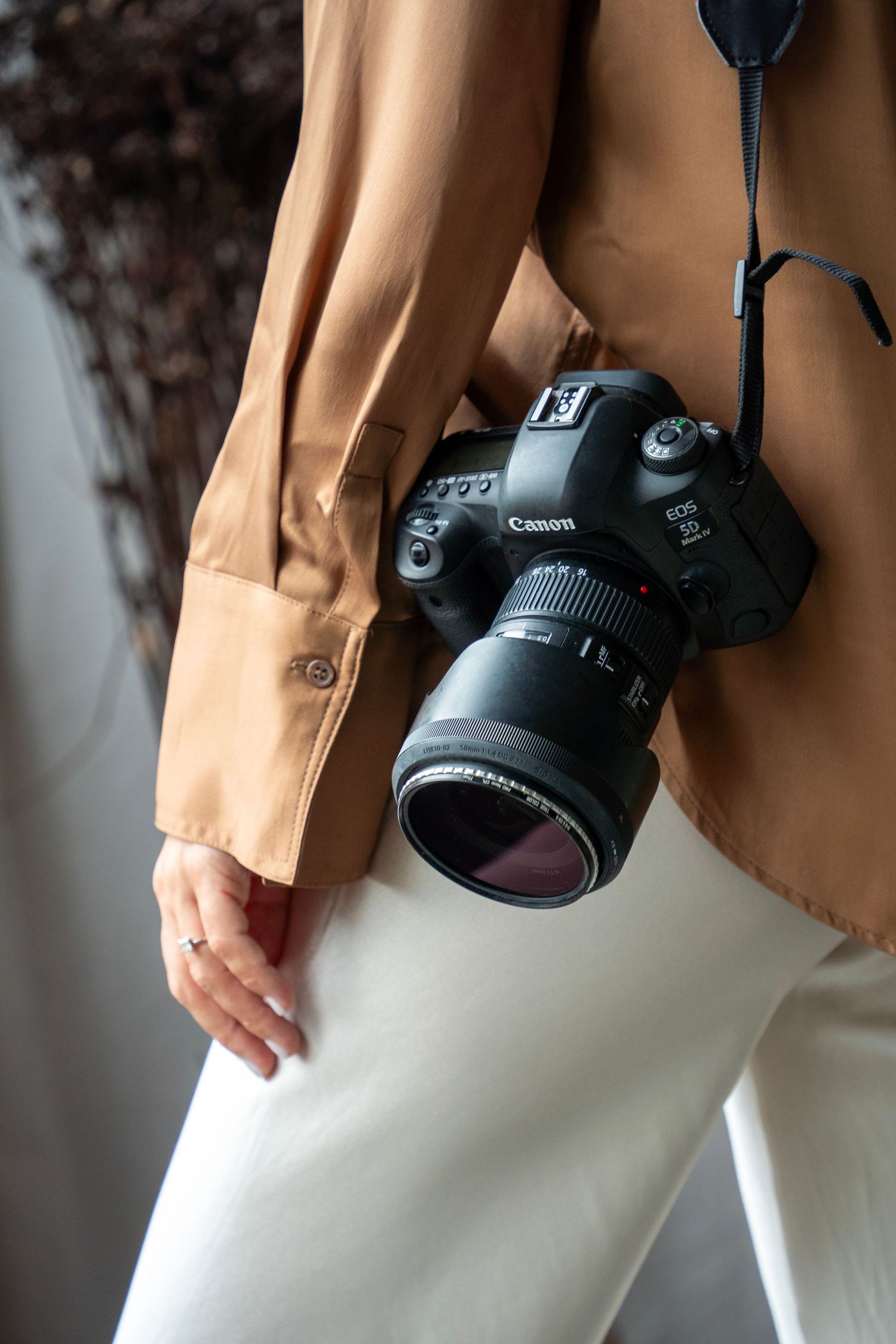 Close-up of a Canon DSLR camera with lens hanging against a beige blazer and white pants.