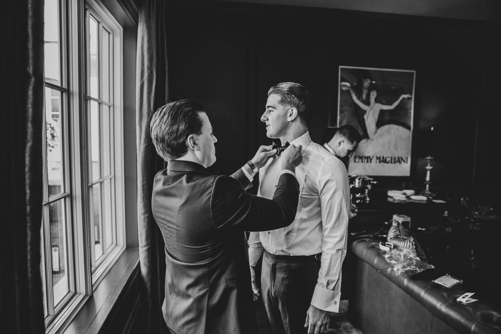 A black and white photograph of wedding preparations by a window as someone adjusts a bowtie.