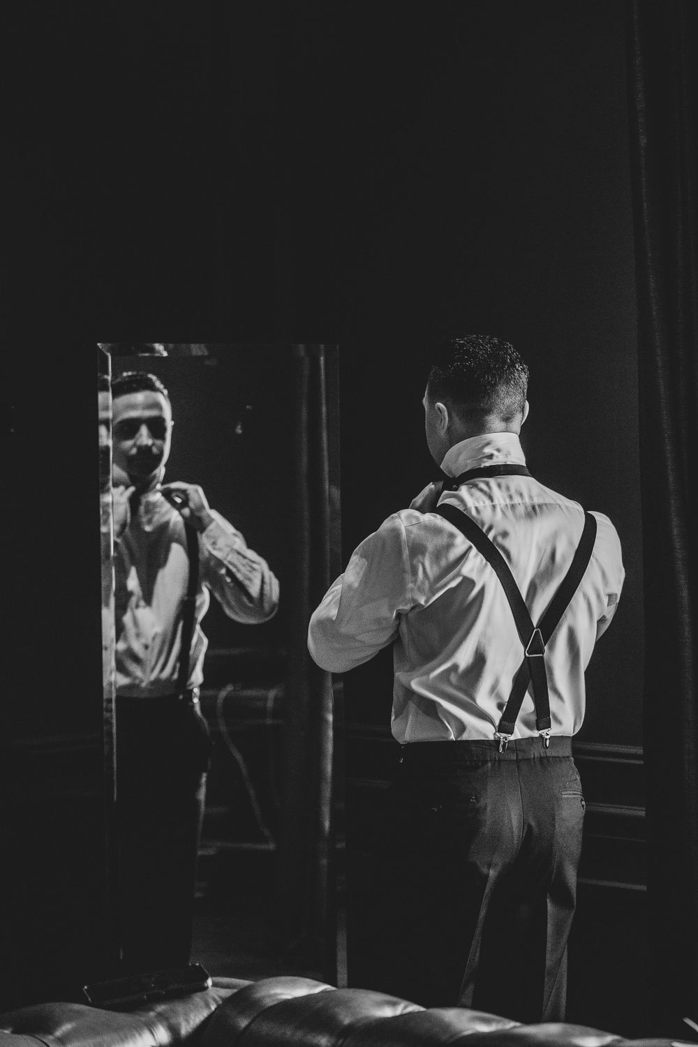 Black and white photo of a person wearing suspenders looking into a mirror in a dark, moody setting.