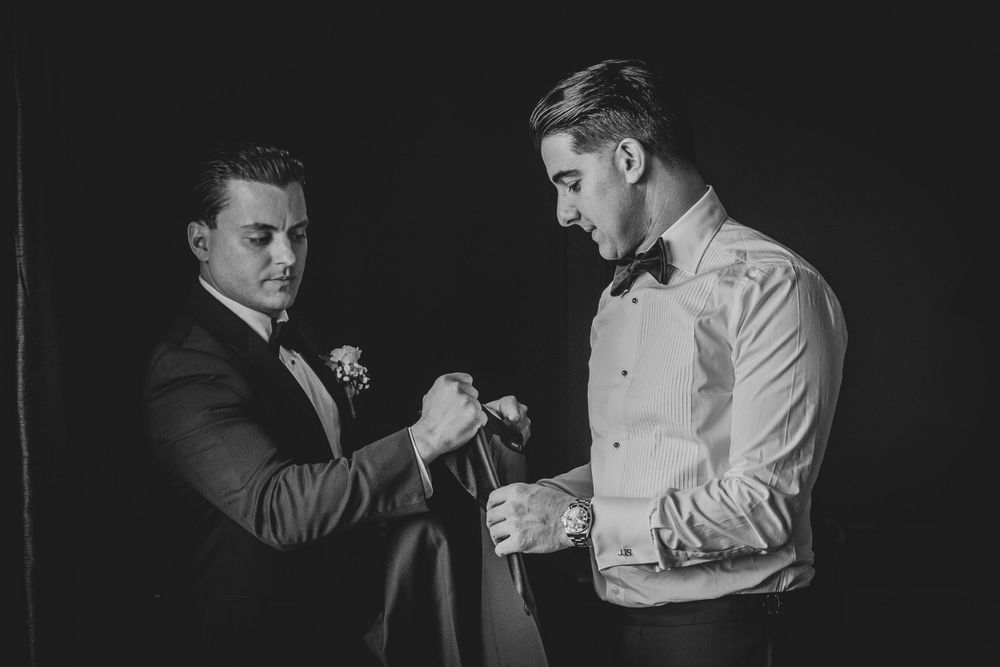 Best man assists with adjusting formal wear and cufflinks in elegant black and white photograph.