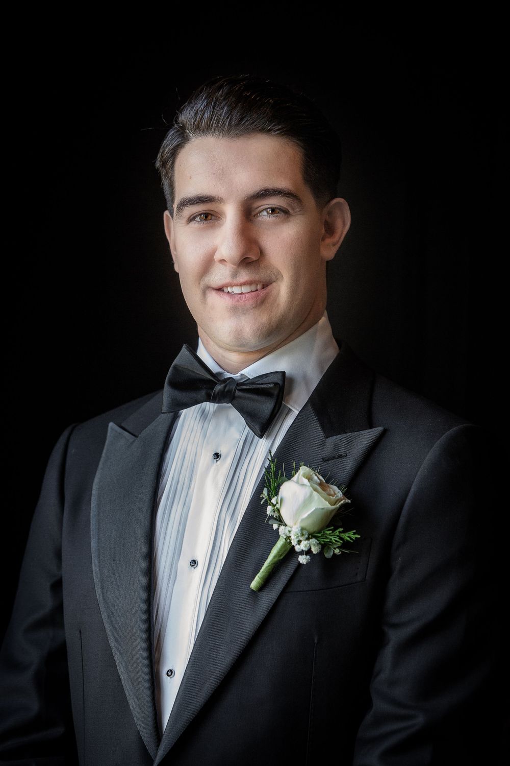 Black and white formal portrait of a groom in a tuxedo with bowtie and white rose boutonniere against dark background.