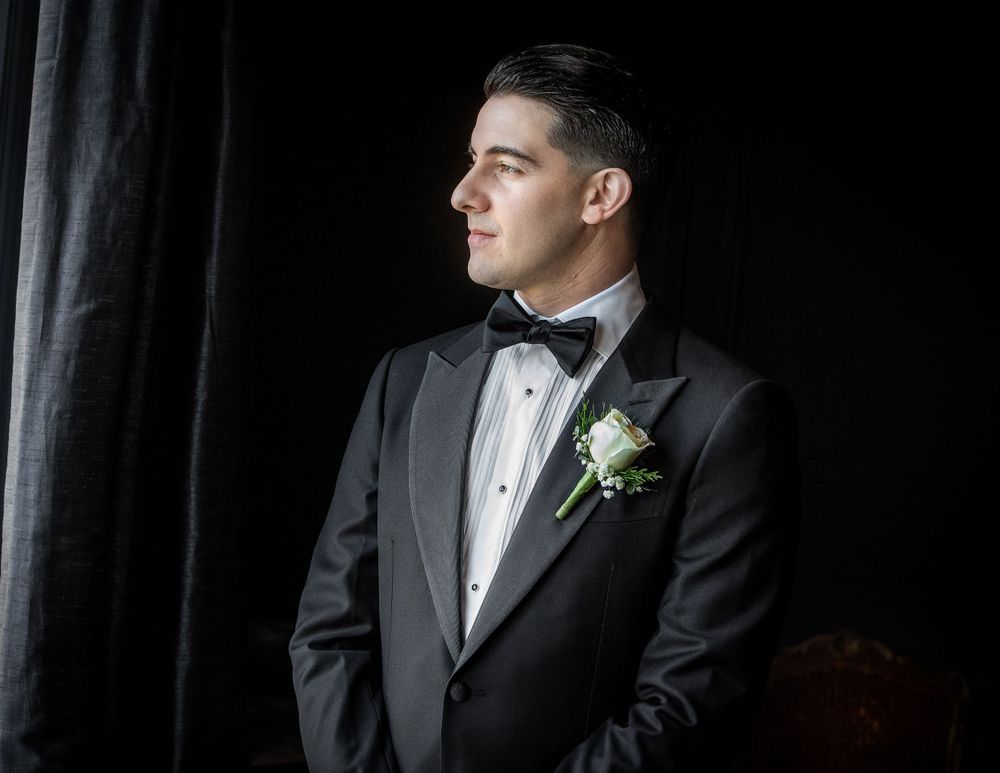 Groom in classic black tuxedo and bow tie with white boutonniere stands in dramatic side lighting against dark background.