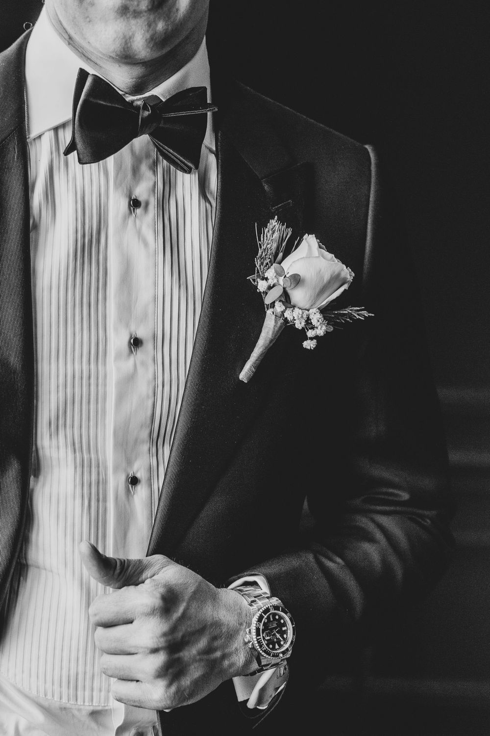 Black and white closeup of formal tuxedo with pleated shirt, bow tie, boutonniere and luxury watch.