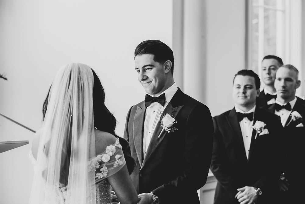 Black and white wedding photo shows couple exchanging joyful glances during ceremony with groomsmen in background.