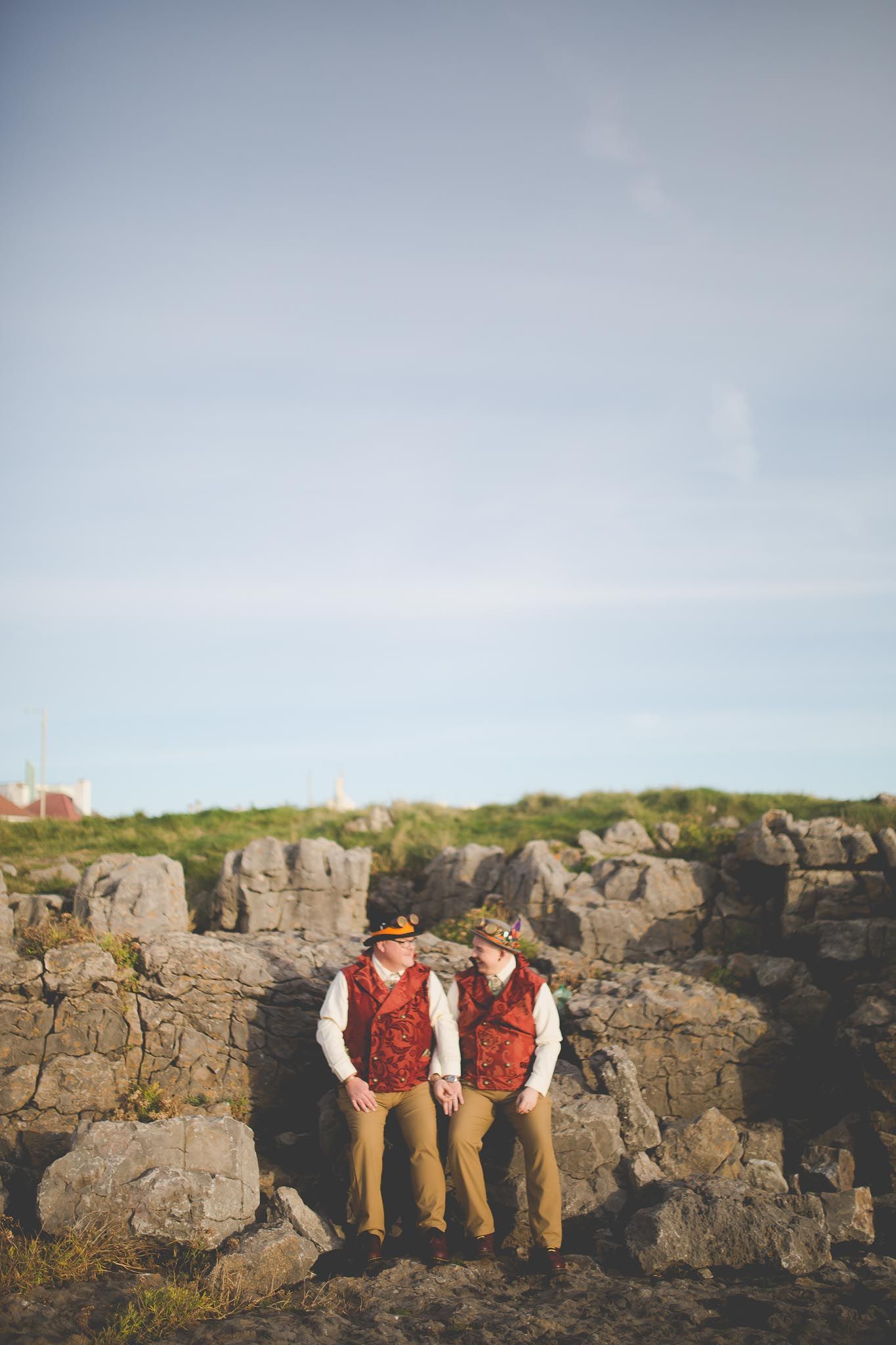 Two people in matching red vests sit together on rocky coastal terrain against a blue sky.