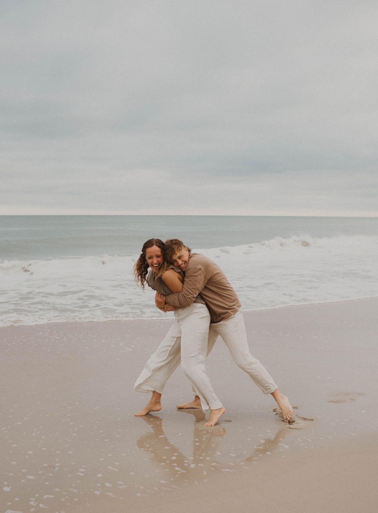 Two people playfully embracing and laughing together on a cloudy beach with waves in the background.