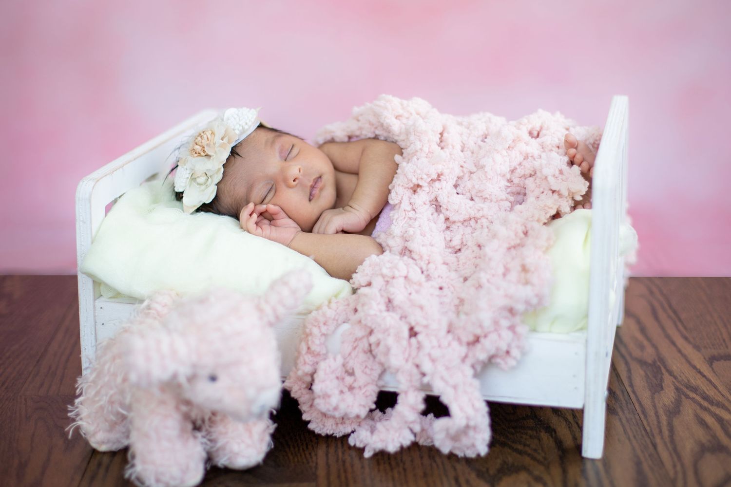 Peaceful sleeping newborn on white bed with pink blanket and plush toys.