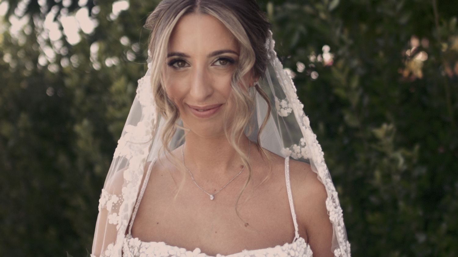 A bride wearing a delicate lace veil smiles at the camera during her wedding day.