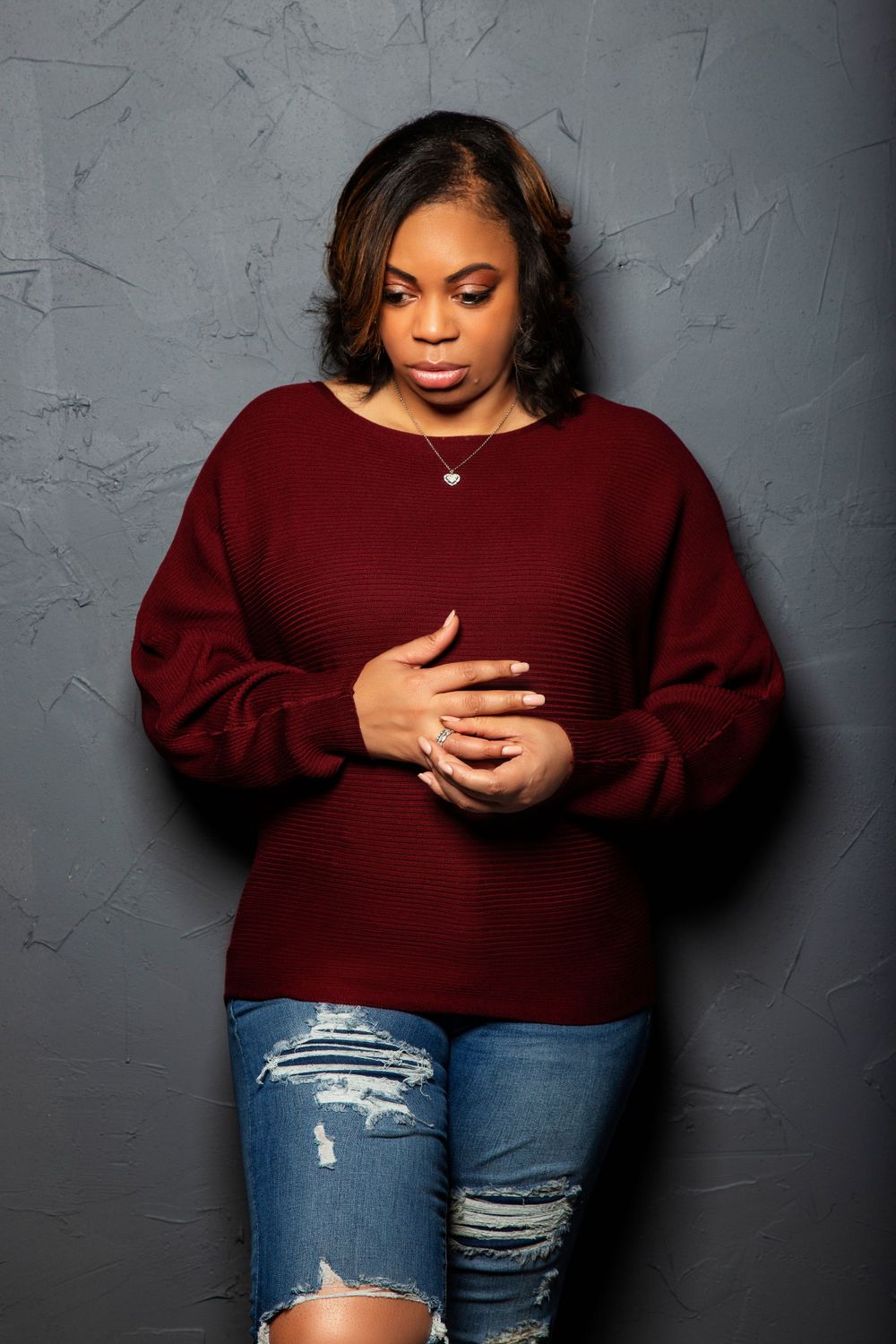 Model in burgundy sweater and distressed jeans poses expressively against grey wall.