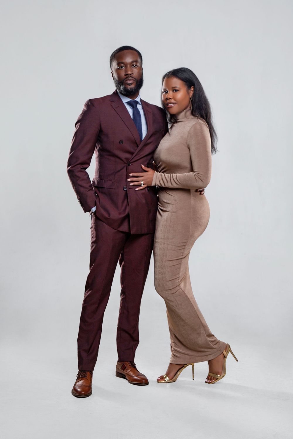 Couple posing in burgundy suit and tan dress against white studio background.