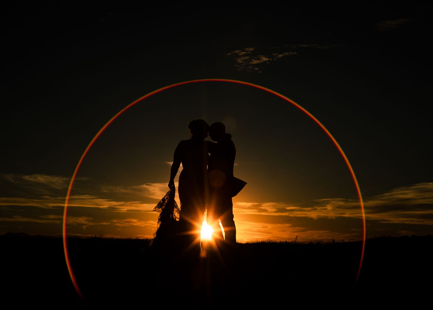 Silhouetted couple stands against a dramatic sunset with a red circular lens flare effect.