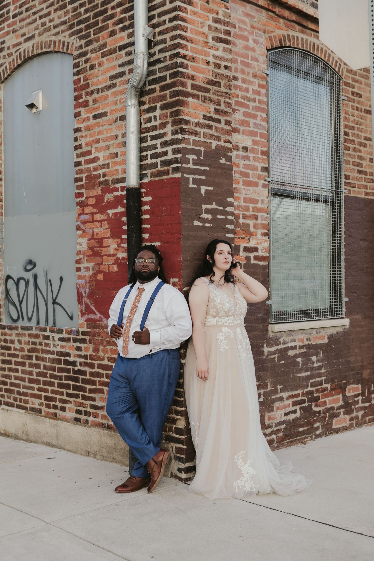 A couple poses stylishly against a brick wall corner wearing formal attire including suspenders and a flowing gown.