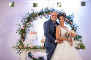 Smiling bride and groom pose together in front of floral arch and wedding cake at reception venue.