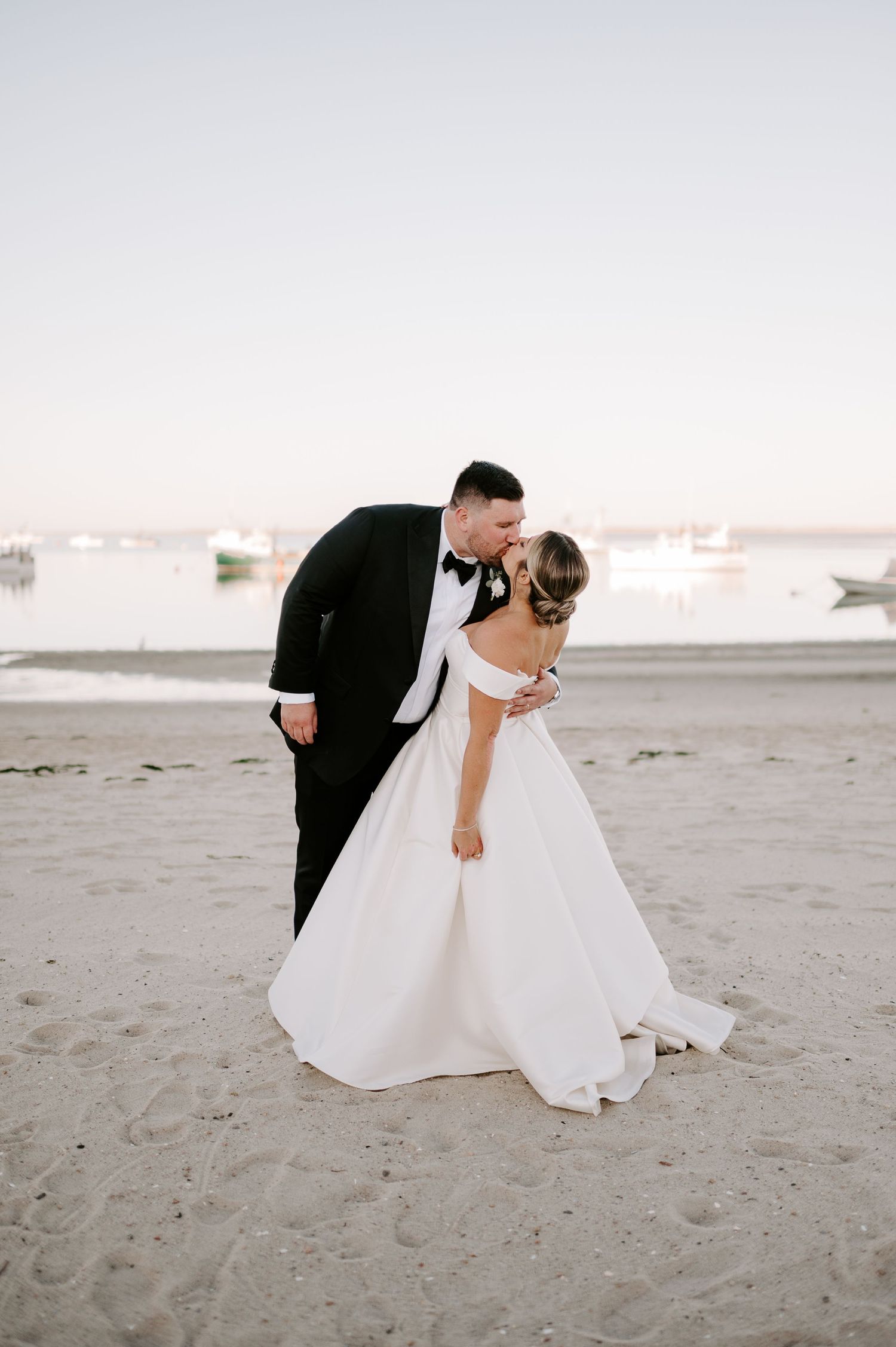 A romantic beach wedding photo sequence showing a couple in formal attire dipping and embracing on the sand at sunset.