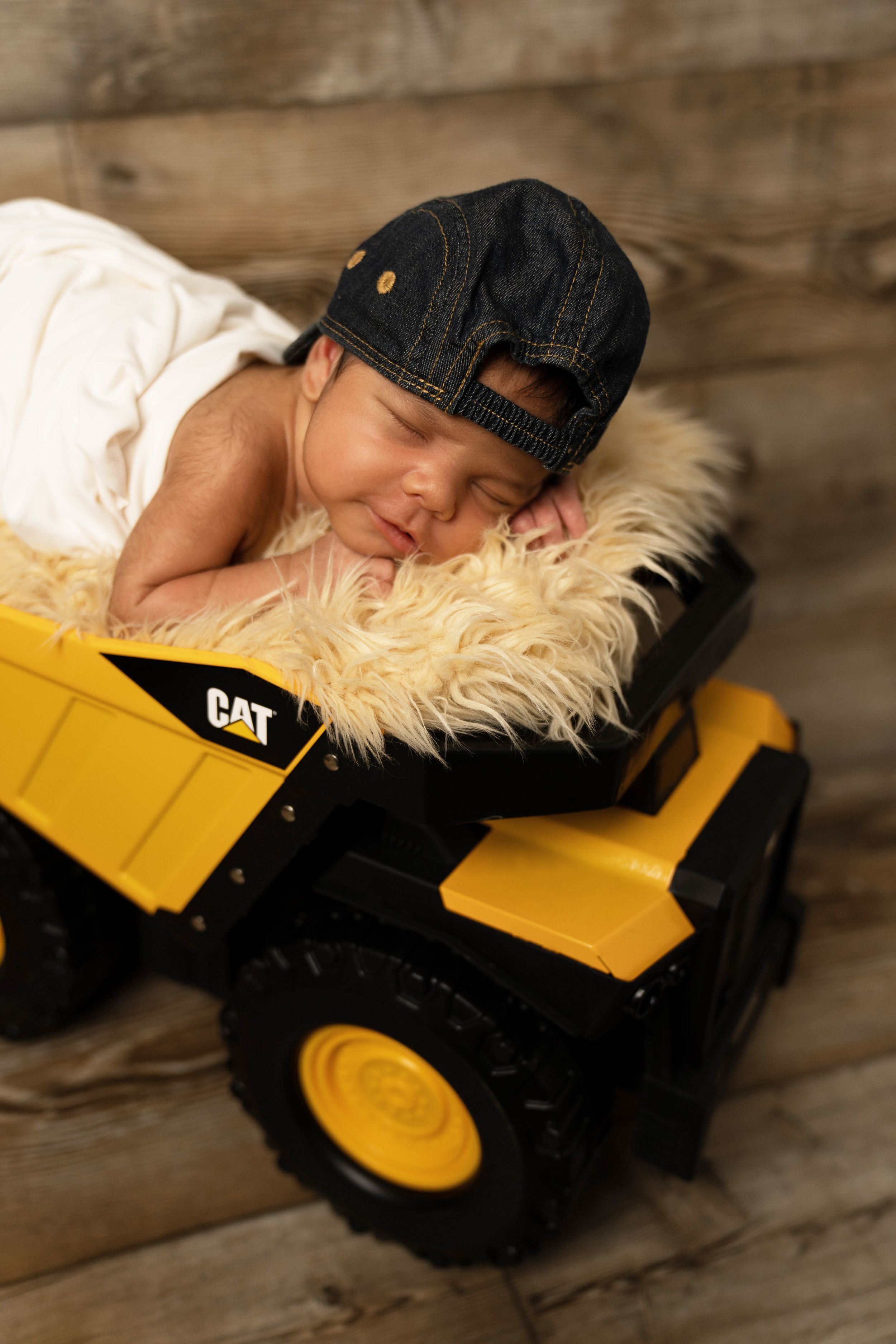Baby sleeping on toy CAT bulldozer wearing denim cap.