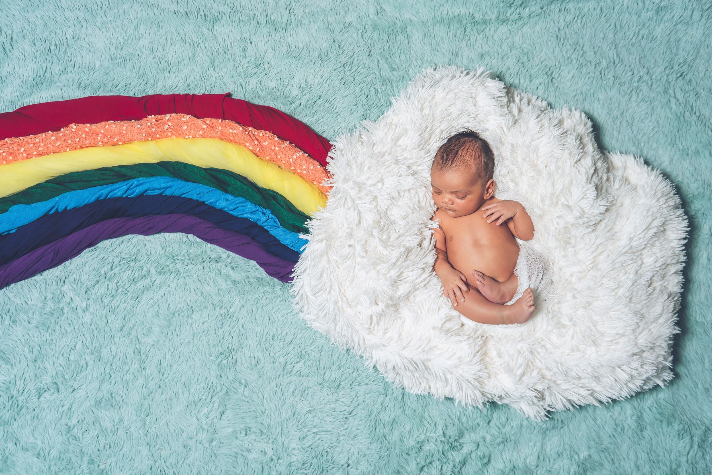 Newborn baby sleeping on white cloud-shaped cushion with rainbow backdrop.