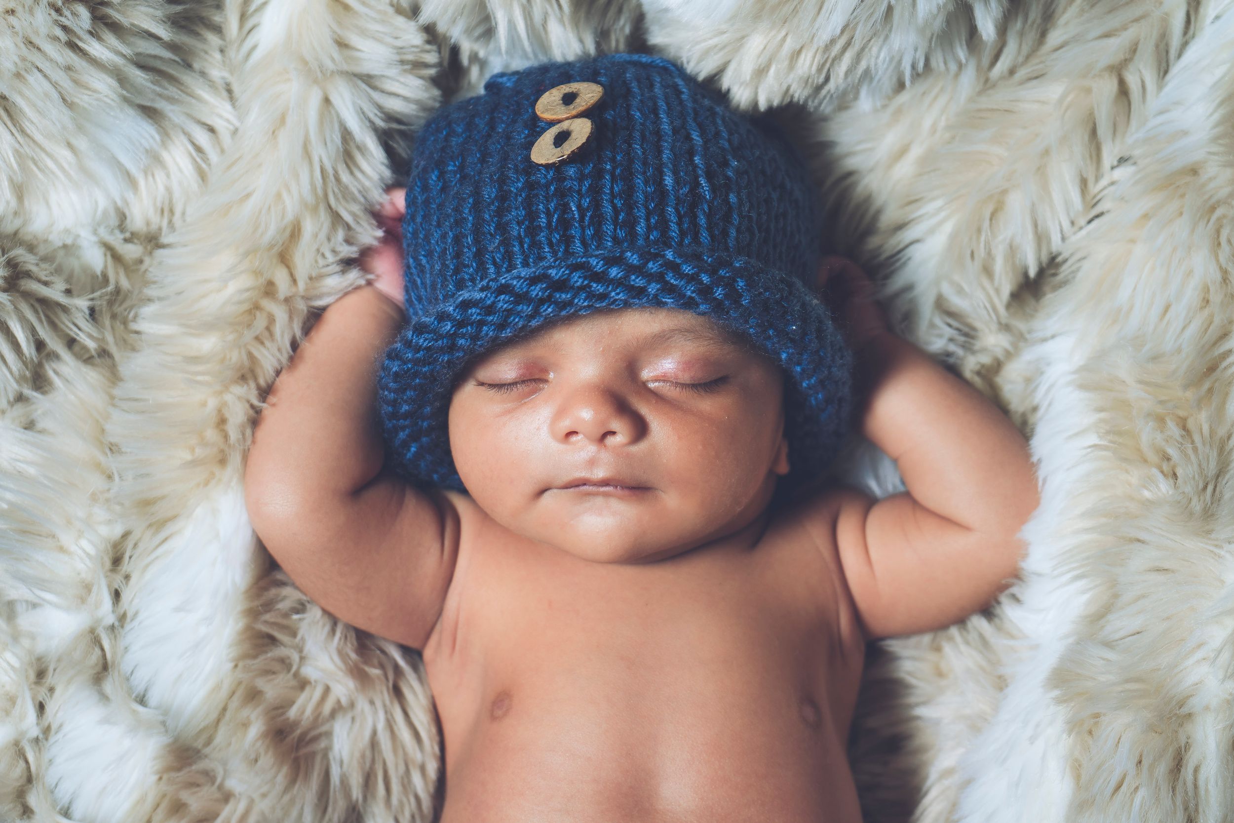 Sleeping newborn wearing knitted blue beanie hat on fuzzy white blanket.