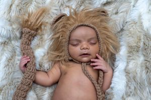 Sleeping newborn wearing lion-themed bonnet on furry backdrop.